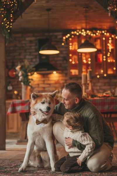 秋田犬とサンタ屋内を待っているニットベージュのセーターで幸せな家族の肖像画 笑顔の両親抱擁とキス彼の小さな息子Eve 12月祝Xmasとともにガーランドライト Noel Tree — ストック写真