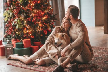 25 Aralık arifesinde evde Akia Inu köpeğiyle kucaklaşan genç bir çiftin portresi. Örgü ören şık elbiseli insanlar, yeni yılı kutlayan şefkat dolu öpücükler, Noel ışıkları, Noel ağacı.