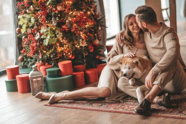 25 Aralık arifesinde evde Akia Inu köpeğiyle kucaklaşan genç bir çiftin portresi. Örgü ören şık elbiseli insanlar, yeni yılı kutlayan şefkat dolu öpücükler, Noel ışıkları, Noel ağacı.