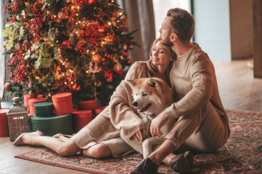 25 Aralık arifesinde evde Akia Inu köpeğiyle kucaklaşan genç bir çiftin portresi. Örgü ören şık elbiseli insanlar, yeni yılı kutlayan şefkat dolu öpücükler, Noel ışıkları, Noel ağacı.
