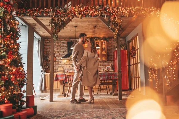 若い素敵なカップルの肖像画屋内の前夜25 12月 家でクリスマスを待っている抱擁のキスを笑って愛好家 エレガントなニットの服の優しさで新年のガーランドライトノエルを祝う — ストック写真