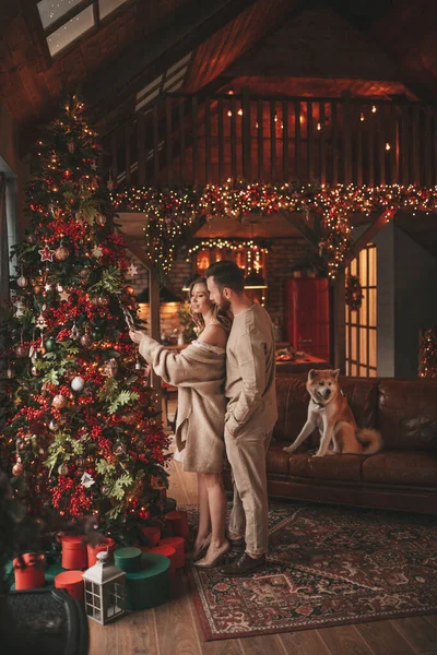 12月25日に秋田犬と一緒に屋内の前夜を抱きしめる若い素敵なカップルの肖像画 ニットスタイリッシュな衣装の人々抱擁キス優しさお祝い新年Garlands Xmasライト家で木 — ストック写真