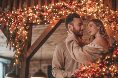 25 Aralık arifesinde evde kucaklaşan güzel bir çiftin portresi. Aşıklar gülüyor, öpücükler evde Noel 'i bekliyor. Yeni yılı kutluyoruz. Çiçek yakıyoruz. Zarif örgü kıyafetlerle süsleniyoruz.