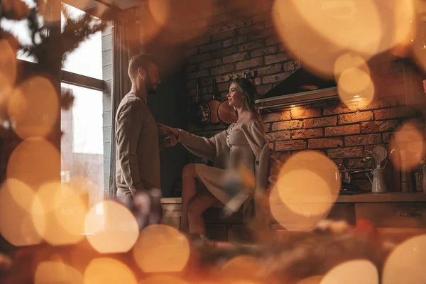 Portrait of young lovely couple hugging indoor eve 25 December. Lovers laughing hugs kisses waiting xmas at home. Celebrating new year garlands lights noel in elegant knitwear outfit tenderness