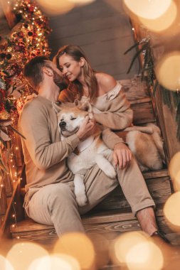 25 Aralık arifesinde evde Akia Inu köpeğiyle kucaklaşan genç bir çiftin portresi. Örgü ören şık elbiseli insanlar, yeni yılı kutlayan şefkat dolu öpücükler, Noel ışıkları, Noel ağacı.