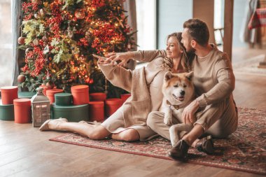 25 Aralık arifesinde evde Akia Inu köpeğiyle kucaklaşan genç bir çiftin portresi. Örgü ören şık elbiseli insanlar, yeni yılı kutlayan şefkat dolu öpücükler, Noel ışıkları, Noel ağacı.