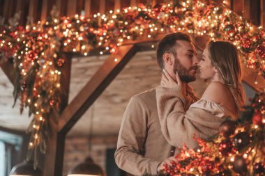 25 Aralık arifesinde evde kucaklaşan güzel bir çiftin portresi. Aşıklar gülüyor, öpücükler evde Noel 'i bekliyor. Yeni yılı kutluyoruz. Çiçek yakıyoruz. Zarif örgü kıyafetlerle süsleniyoruz.