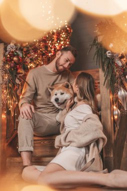 25 Aralık arifesinde evde Akia Inu köpeğiyle kucaklaşan genç bir çiftin portresi. Örgü ören şık elbiseli insanlar, yeni yılı kutlayan şefkat dolu öpücükler, Noel ışıkları, Noel ağacı.