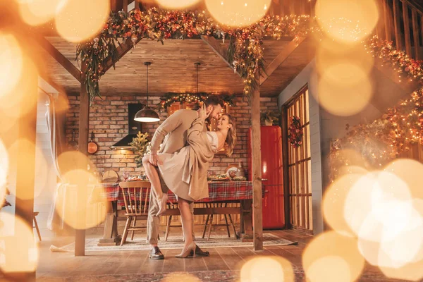 若い素敵なカップルの肖像画屋内の前夜25 12月 家でクリスマスを待っている抱擁のキスを笑って愛好家 エレガントなニットの服の優しさで新年のガーランドライトノエルを祝う — ストック写真