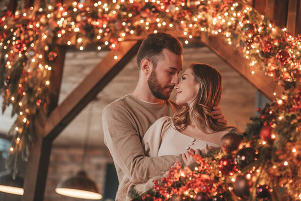 Portrait of young lovely couple hugging indoor eve 25 December. Lovers laughing hugs kisses waiting xmas at home. Celebrating new year garlands lights noel in elegant knitwear outfit tenderness
