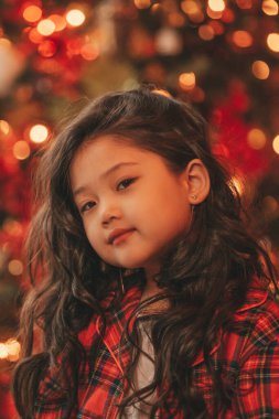 Mutlu küçük Japon hayalperest Noel Baba 'nın Noel ağacının yanında poz vermesini bekliyor. 25 Aralık arifesinde Bokeh xmas 'da yeni yılı kutlayan kırmızı ekose elbiseli Asyalı çocuk.