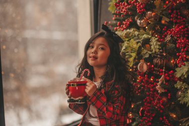 Mucize bekleyen küçük Japon hayalperest kız Noel Baba Noel ağacının yanında şekerleme yiyor. Kırmızı ekoseli Asyalı çocuk yeni yılı kutluyor. 25 Aralık gecesi Bokeh Işıkları 'nda marşmelovlu Xmas bardağı tutuyor.