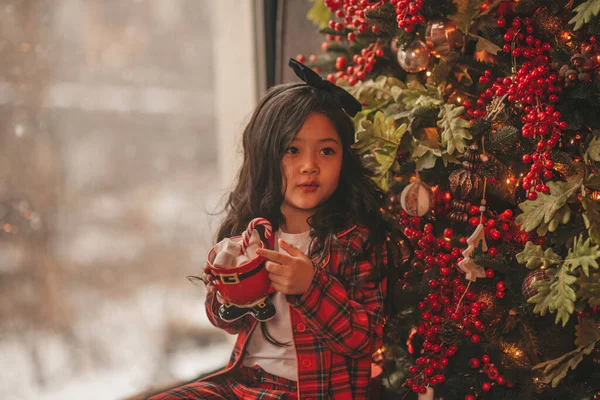 奇跡を待つ幸せな日本の女の子の夢サンタは木の近くでお菓子をお楽しみください アジアの子供で赤い遊び服でお祝いの新年のホールドクリスマスカップとともにマシュマロでBokehライトEve 12月 — ストック写真