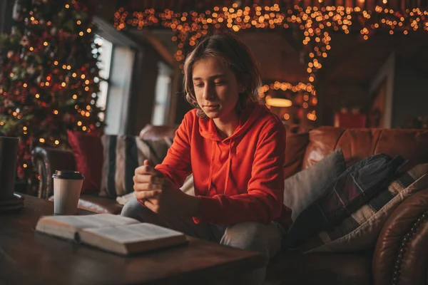 stock image Young teen boy with long hair holding and reading study book for distance learning at home in holidays. Stylish zoomer gen Z celebrates new year with xmas tree bokeh lights garlands eve 25 december