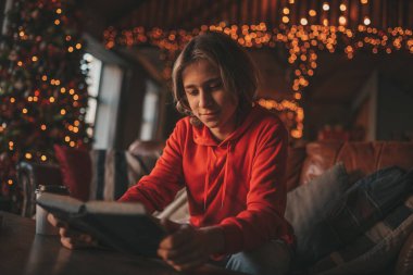 Uzun saçlı genç çocuk tatillerde evden uzakta ders çalışmak için ders kitabı okuyor. Zoomer Z yeni yılı Noel ağacı Bokeh ışıkları ile kutluyor 25 Aralık arifesi