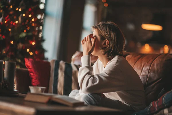 stock image Young teen boy with long hair thoughtful look sad eyes negative mood angry and crying at home. Stylish zoomer gen Z pensive on new year holidays with xmas tree bokeh lights garlands eve 25 december