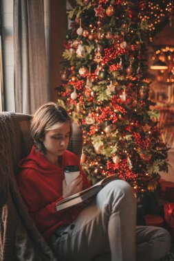 Uzun saçlı genç çocuk tatillerde evden uzakta ders çalışmak için ders kitabı okuyor. Zoomer Z yeni yılı Noel ağacı Bokeh ışıkları ile kutluyor 25 Aralık arifesi