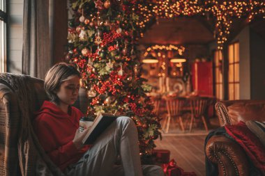Uzun saçlı genç çocuk tatillerde evden uzakta ders çalışmak için ders kitabı okuyor. Zoomer Z yeni yılı Noel ağacı Bokeh ışıkları ile kutluyor 25 Aralık arifesi