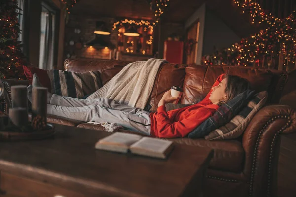 長い髪の読書の勉強書を持つ若い10代の男の子と自宅で熱い飲み物の距離学習をお楽しみください スタイリッシュなZoomer Gen Zは12月25日を祝うクリスマスツリーボケライトガーランド — ストック写真