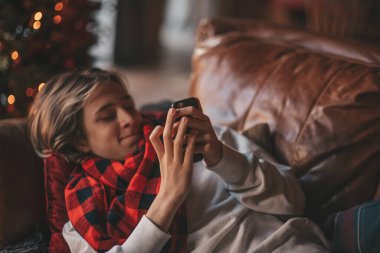 Uzun saçlı, akıllı telefon tutan genç bir çocuk. Mesajlaşmak için kablosuz teknoloji mobil cihaz kullanıyor. Zarif çocuk jenerasyonu Z, 25 Aralık arifesinde Noel 'i Noel ağacı ışıklarıyla kutluyor.