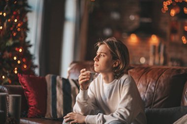 Uzun saçlı, rahat ve düşünceli genç bir çocuk. Evde sıcak içkinin tadını çıkarıyor. Zoomer gen Z yeni yıl tatilini Noel ağacıyla kutluyor. Bokeh ışıkları çelenkleri 25 Aralık arifesi.