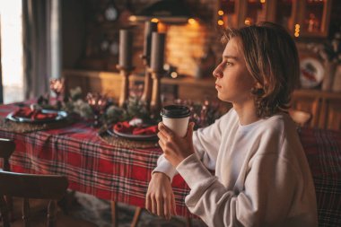 Uzun saçlı, rahat ve düşünceli genç bir çocuk. Evde sıcak içkinin tadını çıkarıyor. Zoomer gen Z yeni yıl tatilini Noel ağacıyla kutluyor. Bokeh ışıkları çelenkleri 25 Aralık arifesi.