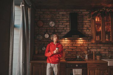 Uzun saçlı, rahat ve düşünceli genç bir çocuk. Evde sıcak içkinin tadını çıkarıyor. Zoomer gen Z yeni yıl tatilini Noel ağacıyla kutluyor. Bokeh ışıkları çelenkleri 25 Aralık arifesi.