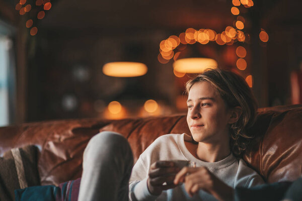 Молодой эмоциональный веселый подросток с длинными волосами смеется и радуется в уютном доме. Стильный зоопарк ген Z в хорошем настроении празднует Новый год с рождественской елкой боке огней гирлянды накануне 25 декабря