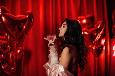 Sensual curly black haired woman in fishnet gloves with glass of wine posing on red glamour background 