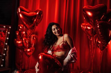 Young tempting brunette woman with perfect skin in sexy lingerie posing with heart balloons in a red room  