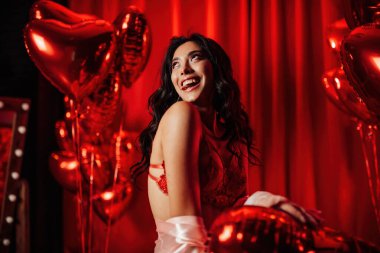 Young tempting brunette woman with perfect skin in sexy lingerie posing with heart balloons in a red room  
