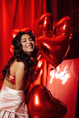 Young tempting brunette woman with perfect skin in sexy lingerie posing with heart balloons in a red room  