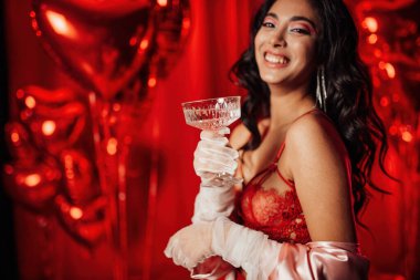 Sensual curly black haired woman in fishnet gloves with glass of wine posing on red glamour background 