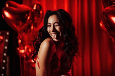 Young tempting brunette woman in sexy lingerie shows her tongue and posing with red heart balloons in studio 