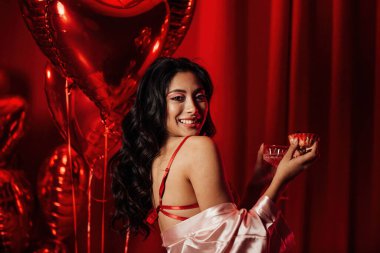 Sensual young japanese black hair woman in sexy lingerie posing with cupcake and coupe glass of champagne at red glamour background with balloons