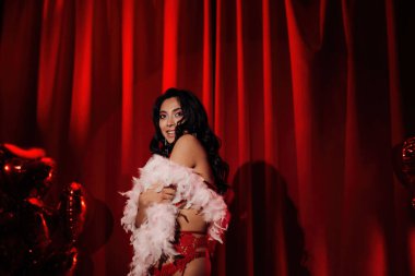 Sensual curly black hair asian woman with feather boa posing on red glamour background