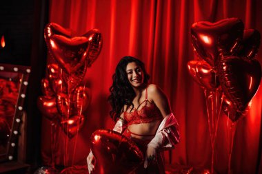 Young tempting brunette woman with perfect skin in sexy lingerie posing with heart balloons in a red room  