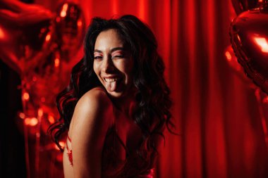 Young tempting brunette woman in sexy lingerie shows her tongue and posing with red heart balloons in studio 