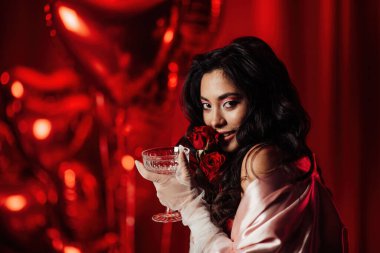 Sensual curly black haired woman in fishnet gloves with glass of wine and roses posing on red glamour background 