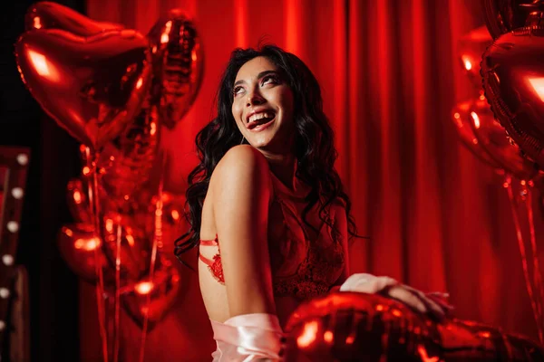 Young tempting brunette woman with perfect skin in sexy lingerie posing with heart balloons in a red room  