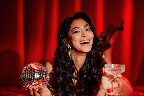 Curly black hair asian model with disco ball and champagne glass posing in bed on Valentine\'s day