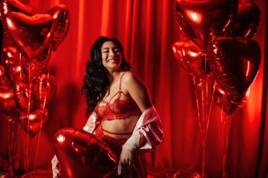 Young tempting brunette woman with perfect skin in sexy lingerie posing with heart balloons in a red room  