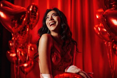 Young tempting brunette woman with perfect skin in sexy lingerie posing with heart balloons in a red room  