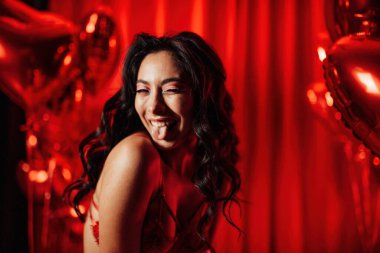 Young tempting brunette woman in sexy lingerie shows her tongue and posing with red heart balloons in studio 