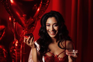 Sensual young japanese black hair woman in sexy lingerie posing with cupcake and coupe glass of champagne at red glamour background with balloons