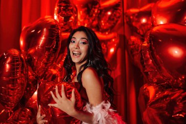 Young tempting brunette woman in sexy lingerie shows her tongue and posing with red heart balloons in studio 