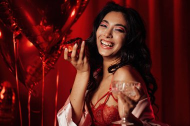 Sensual young japanese black hair woman in sexy lingerie posing with cupcake and coupe glass of champagne at red glamour background with balloons