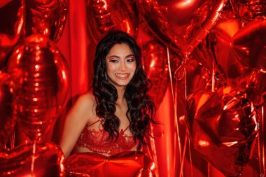 Young tempting brunette woman with perfect skin in sexy lingerie posing with heart balloons in a red room  