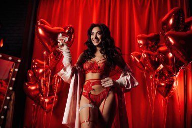 Sensual curly black haired woman in fishnet gloves with glass of wine posing on red glamour background 