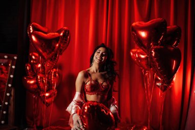 Young tempting brunette woman with perfect skin in sexy lingerie posing with heart balloons in a red room  